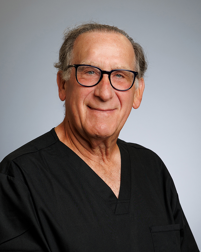 The image shows a man wearing glasses, smiling, and posing for the photograph. He is dressed in a black shirt and has gray hair.