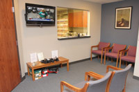 The image shows an interior space that appears to be a waiting room or reception area within a medical facility, with a television mounted on the wall above a display case, chairs arranged for seating, and a countertop with information materials.