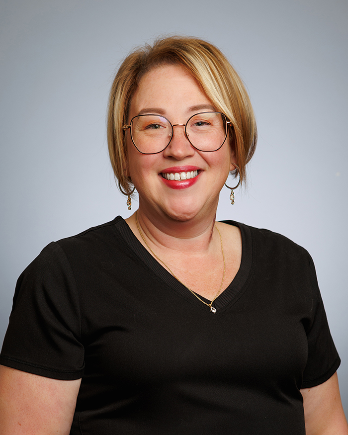 The image is a portrait of a woman smiling at the camera. She has short hair, wears glasses and a black top with a necklace.