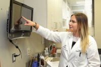 A woman in a white lab coat is pointing at something on a television screen.